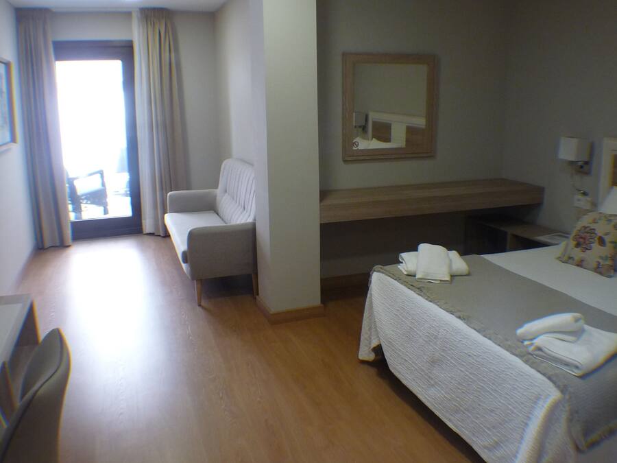 Habitación de hotel con cama, escritorio, silla, mesita, espejo y ventana con cortinas.