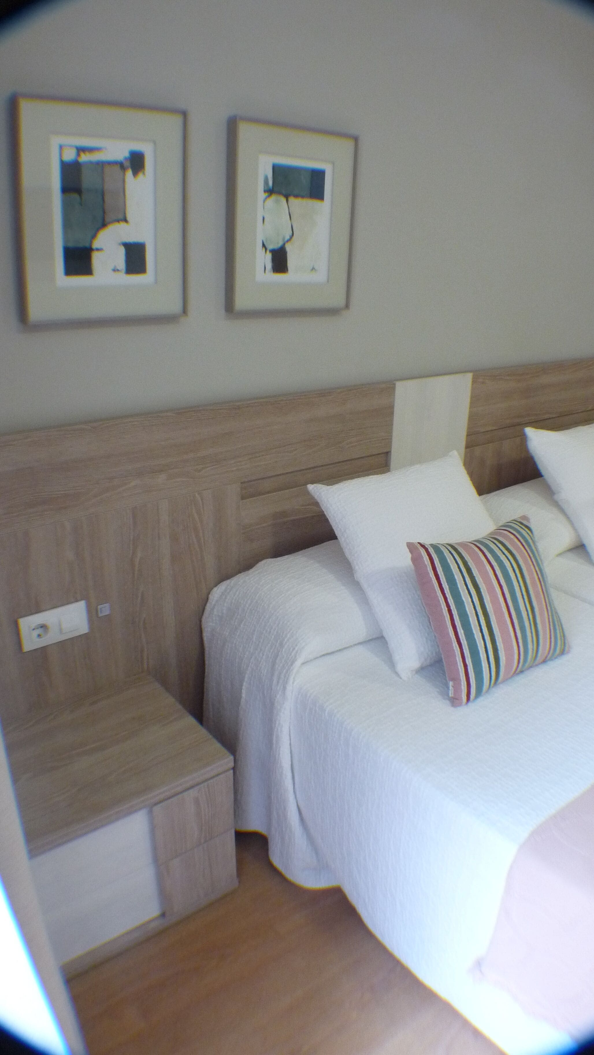 Un dormitorio con cabecera de madera, dos camas con ropa de cama blanca, un almohadón a rayas y dos cuadros enmarcados en la pared.