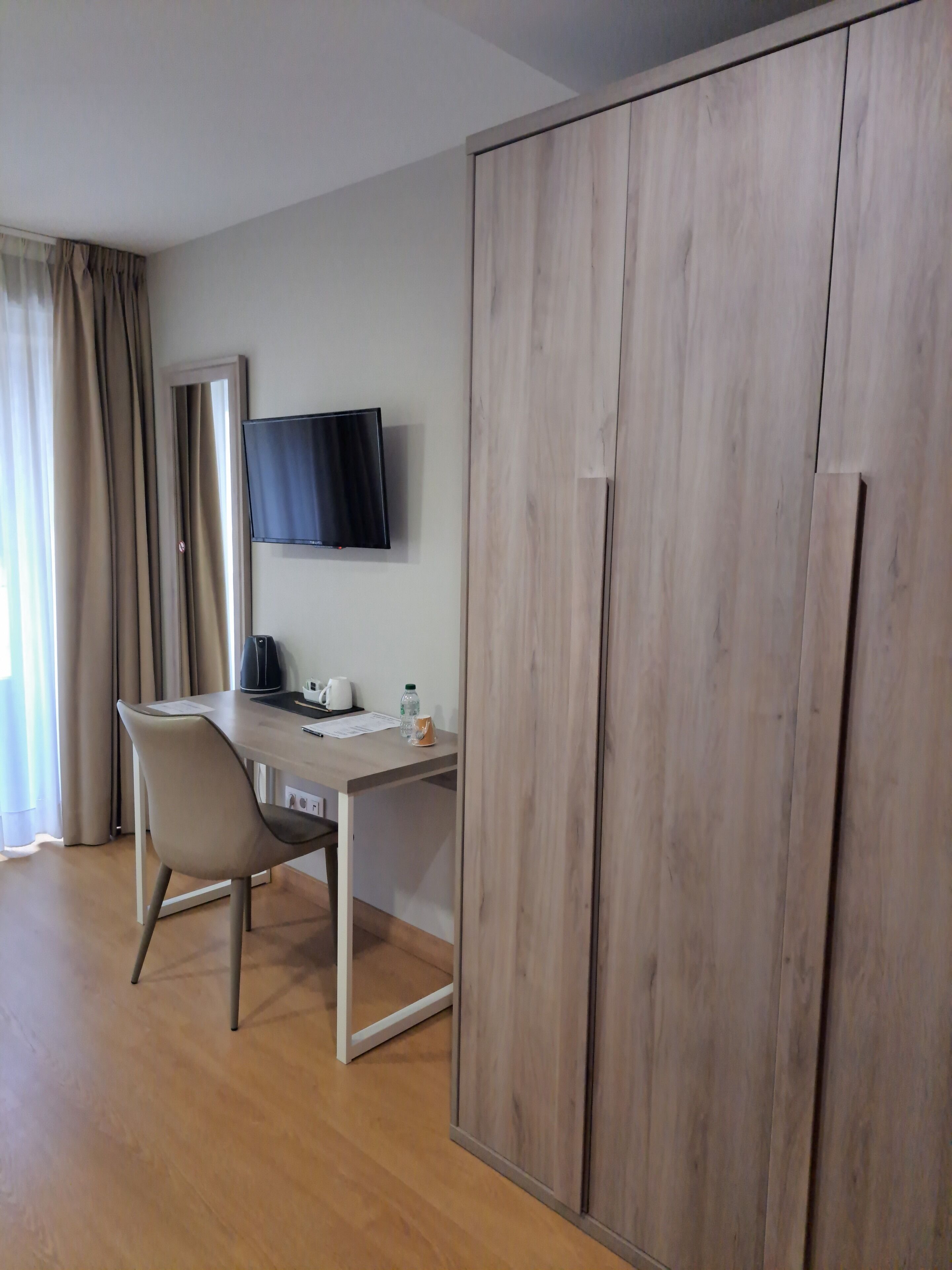 A room with a wooden wardrobe, a desk with a chair, a flat-screen TV, and a window with curtains.