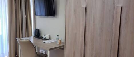 A room with a wooden wardrobe, a desk with a chair, a flat-screen TV, and a window with curtains.
