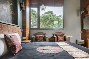 Una habitación con una cama, dos sillas de madera, una ventana con vista a árboles y una lámpara colgante.