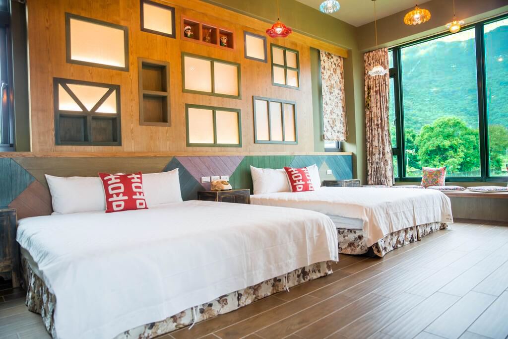 A hotel room with two beds, a large window with a view of greenery, and a wall with geometric patterns.