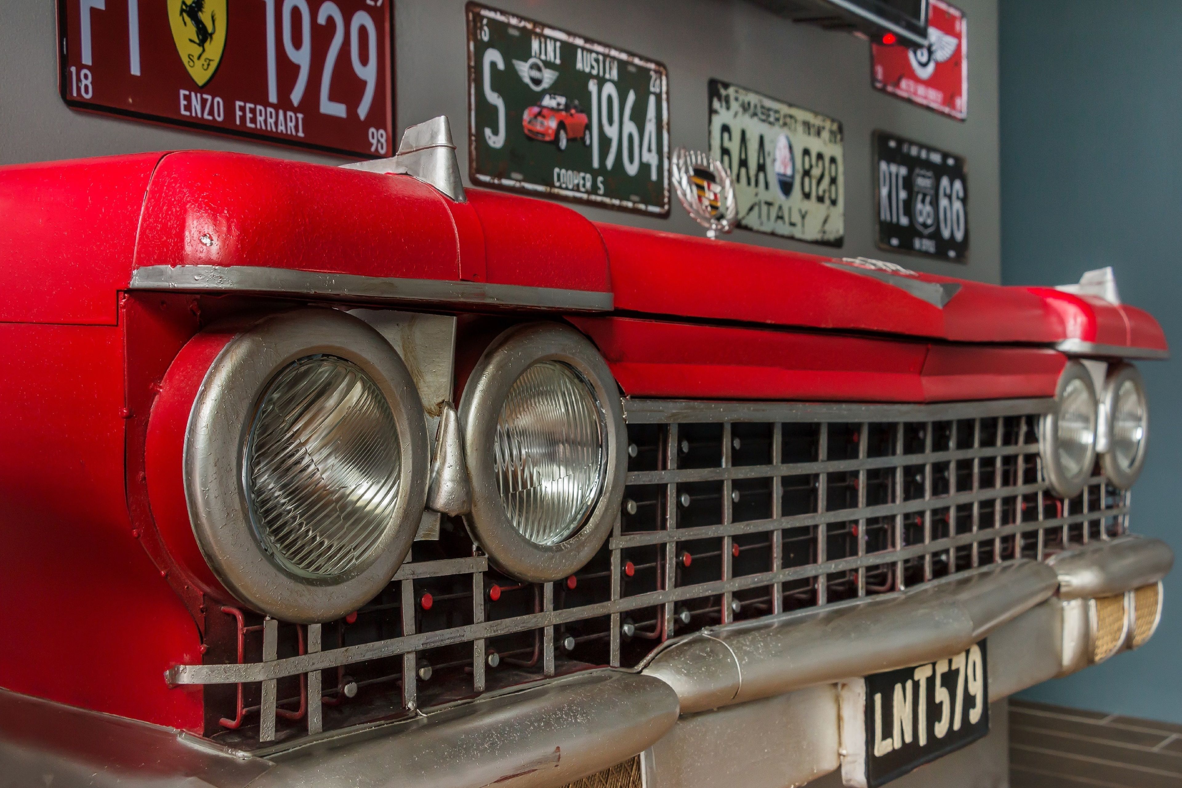Un coche antiguo con una prominente parrilla frontal y faros, y varias placas de matrícula visibles en la pared de fondo.