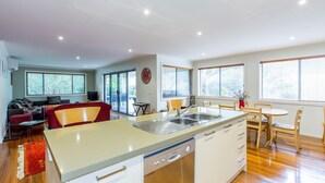 Una cocina moderna con una isla central, un comedor con sillas de madera, un sofá y un televisor en una sala de estar con ventanas amplias.