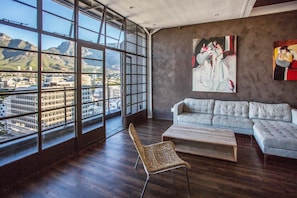 A modern living room with a large window, a sofa, a chair, and a painting. - Manhattan Penthouse (Cape Town)