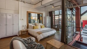 Une chambre moderne avec un grand lit, une table de chevet en bois, un bureau et une vue sur la ville grâce à de grandes fenêtres.