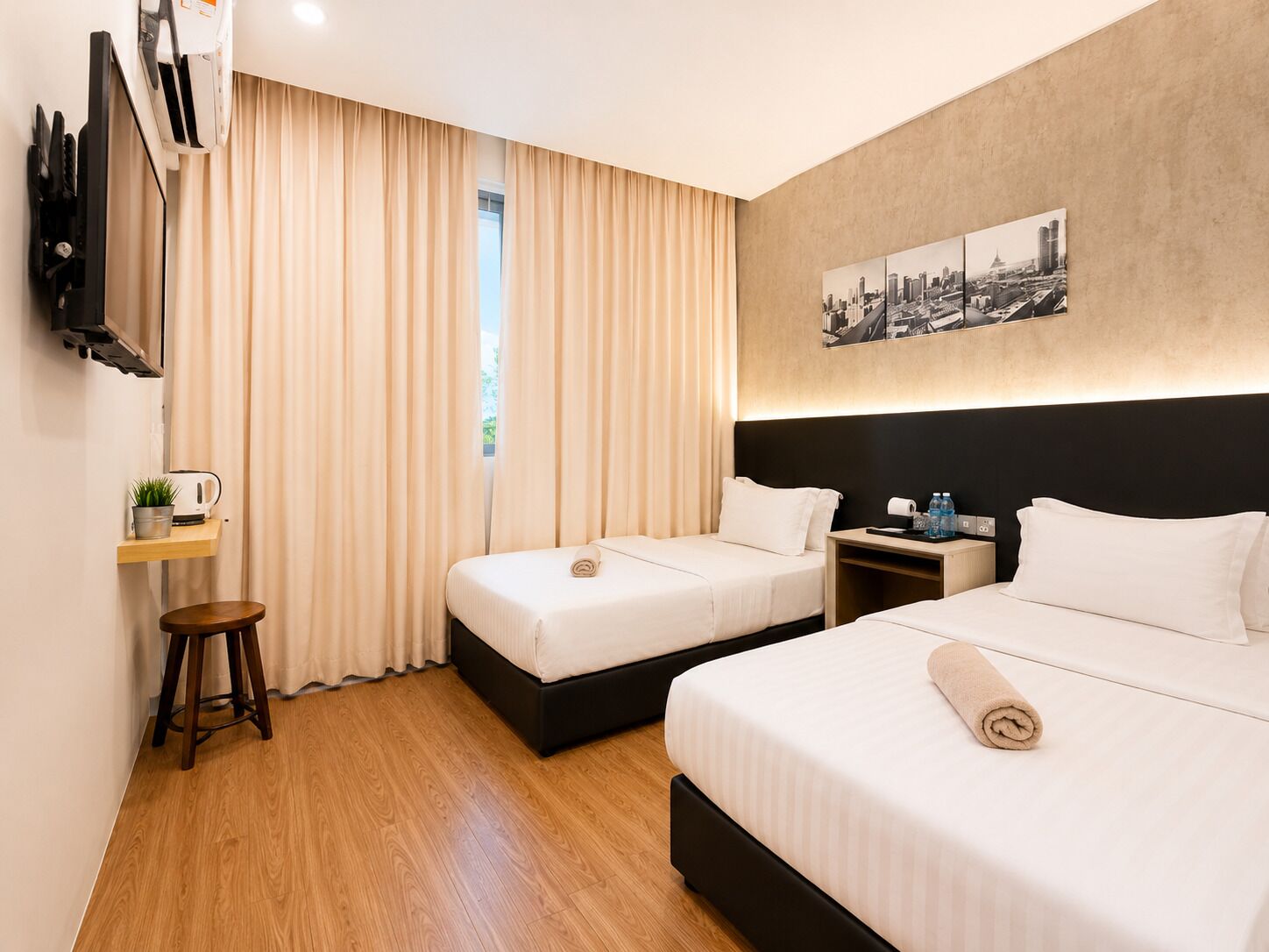 Une chambre d’hôtel avec deux lits, une télévision, une petite table avec une bouilloire, une plante et un tableau représentant un paysage urbain accroché au mur.