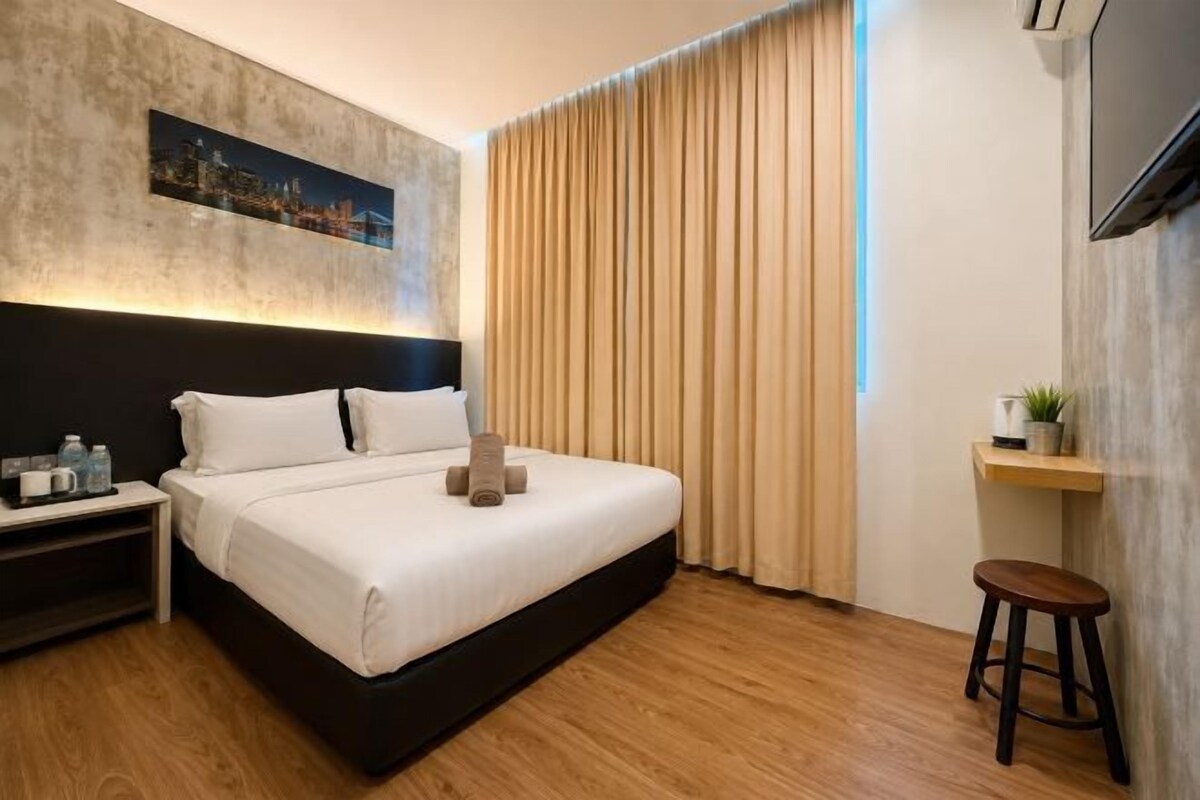 Een moderne hotelkamer met een bed, nachtkastje, televisie, raam met gordijnen en een klein krukje.