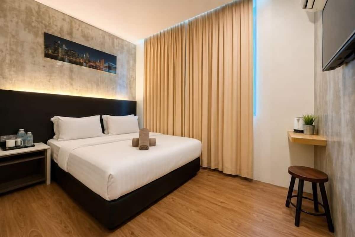 Ein modernes Hotelzimmer mit Bett, Nachttisch, Fernseher, einem Fenster mit Vorhängen und einem kleinen Hocker.