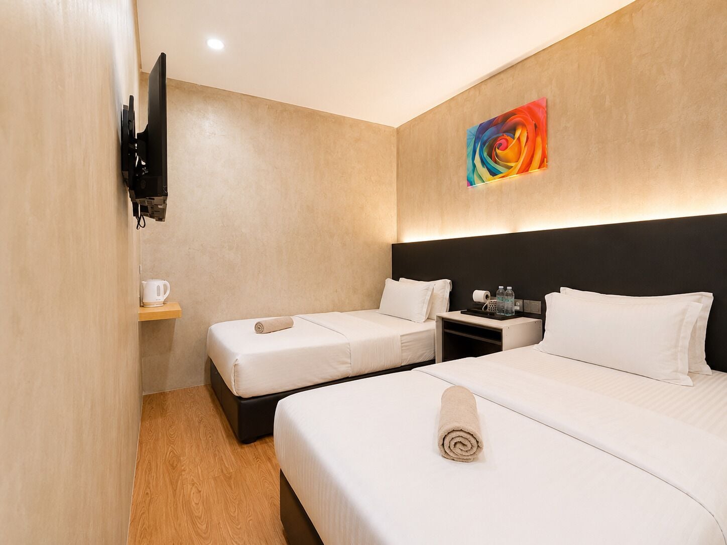 A hotel room with two beds, a flat-screen TV mounted on the wall, a painting, and a small table with a kettle and water bottles.