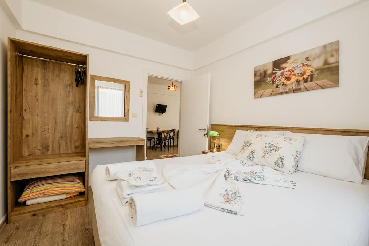 A neatly arranged bedroom with a wooden wardrobe, a bed with floral bedding, a painting on the wall, and a small table with a lamp.