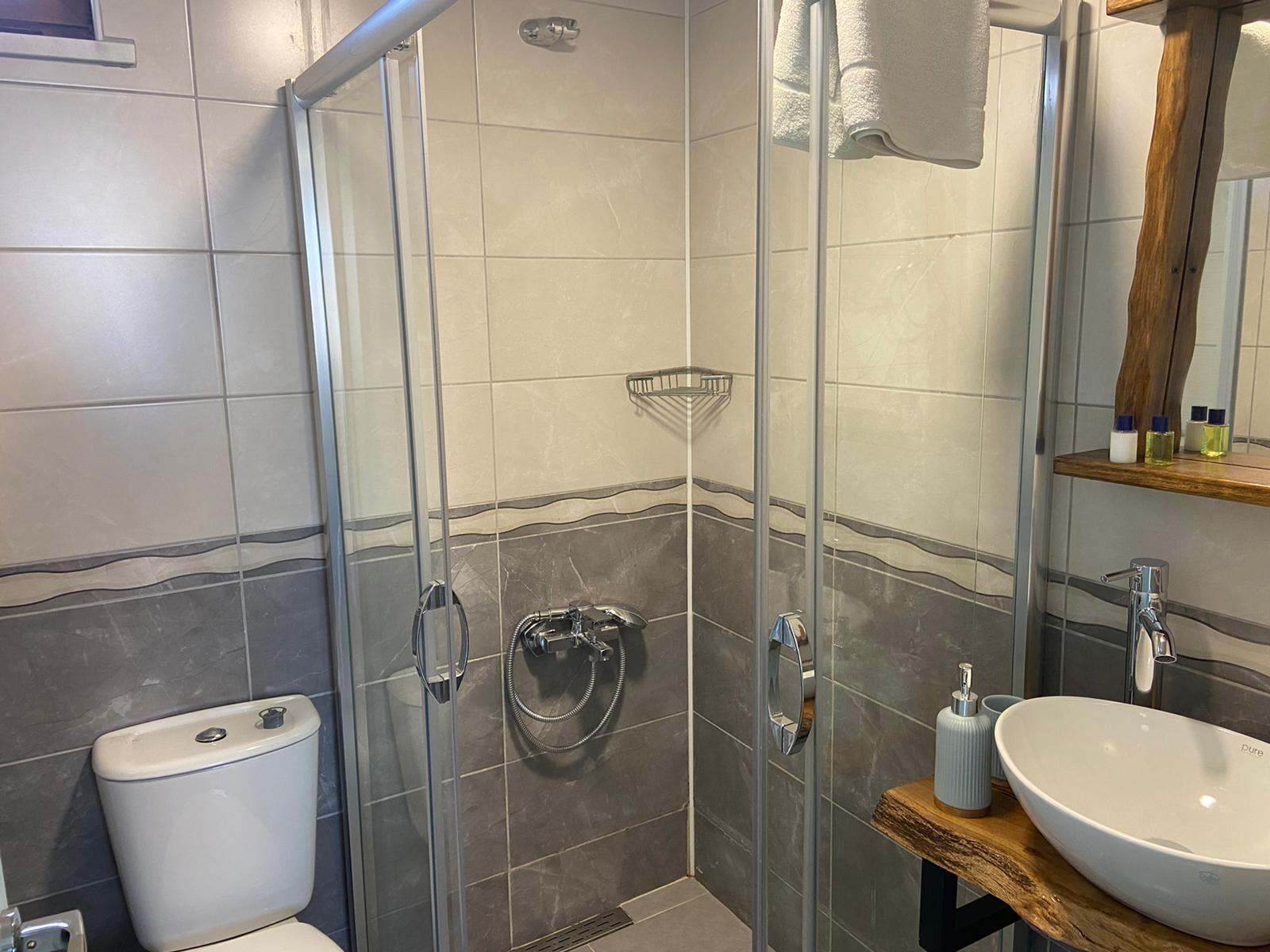 Un baño moderno con ducha envuelta en vidrio, inodoro blanco y tocador de madera con un lavamanos grande.