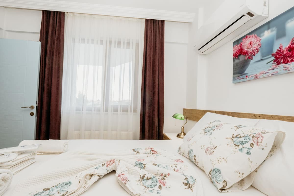 A hotel room with a bed, a nightstand with a lamp, a painting of flowers on the wall, and a window with curtains.