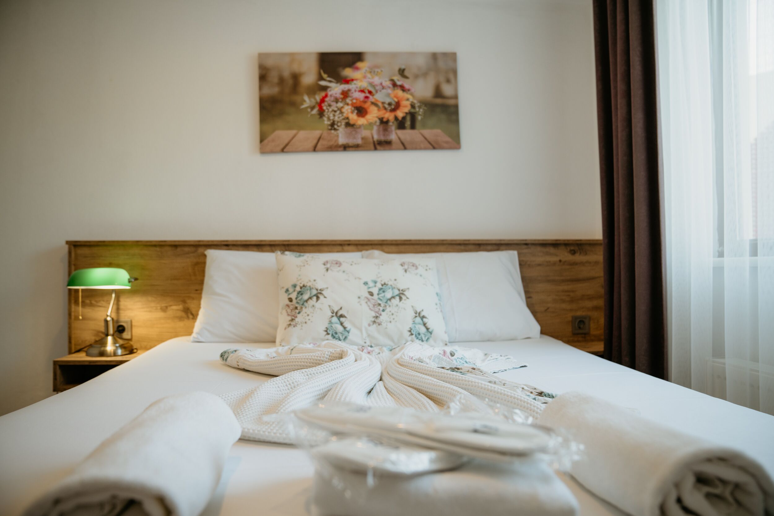 A neatly made bed with a floral pillow, a green lamp on a wooden nightstand, and a painting of flowers on the wall.