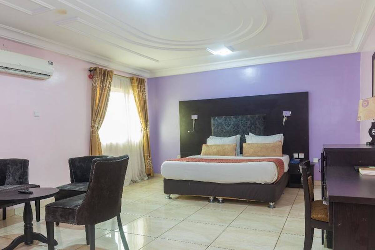 A hotel room with a bed, a desk, two chairs, a window with curtains, and an air conditioning unit.