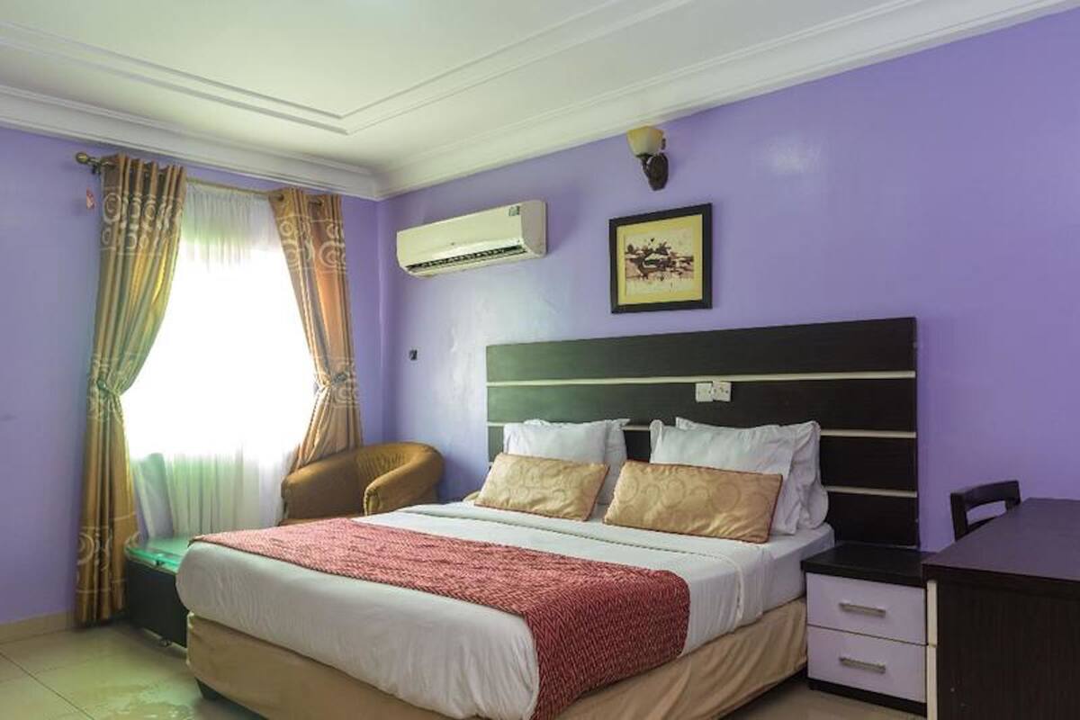 A bedroom with a bed, a chair, a desk, an air conditioner, and a framed picture on the wall.