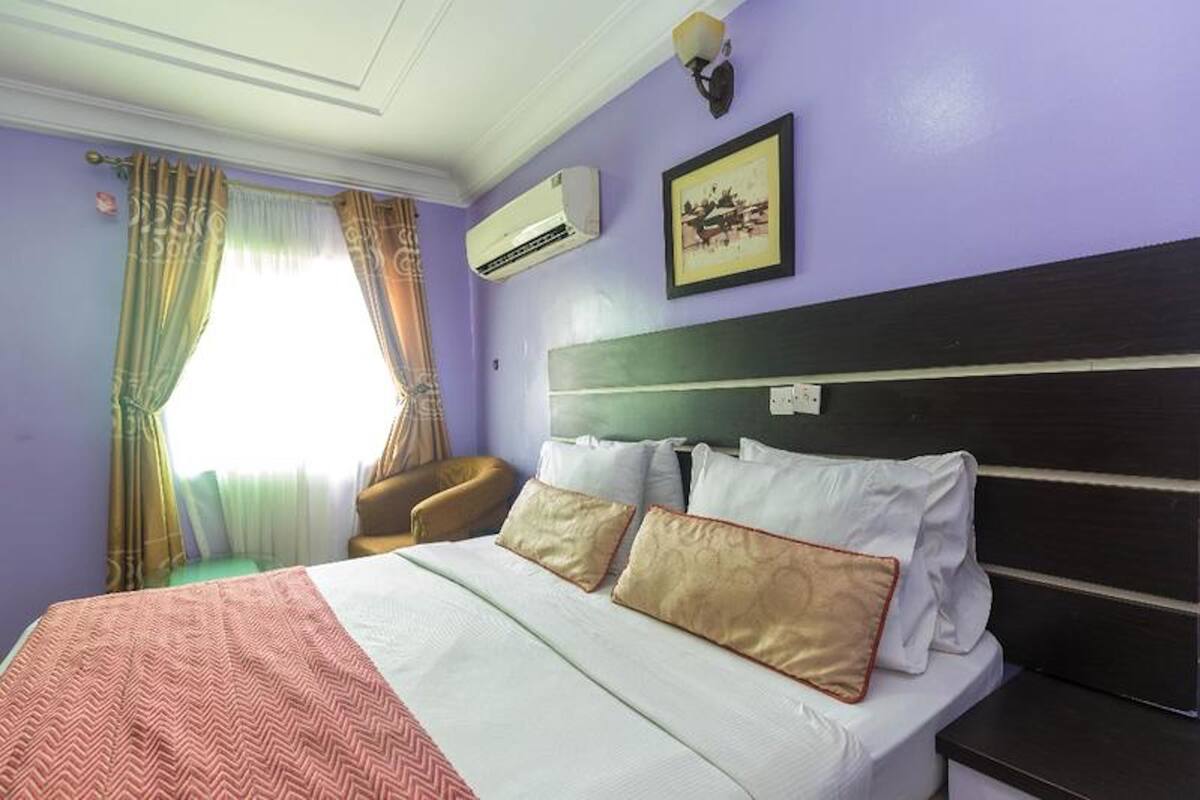 A hotel room with a bed, a chair, an air conditioning unit, and a framed picture on the wall.