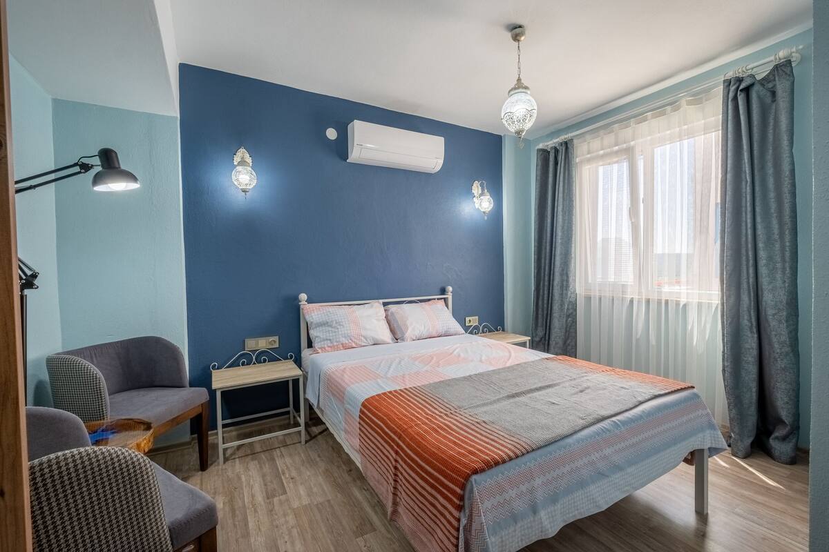 Ein Schlafzimmer mit einem Bett, zwei Nachttischen, einer Lampe, einem Sessel und einem Fenster mit Vorhängen.