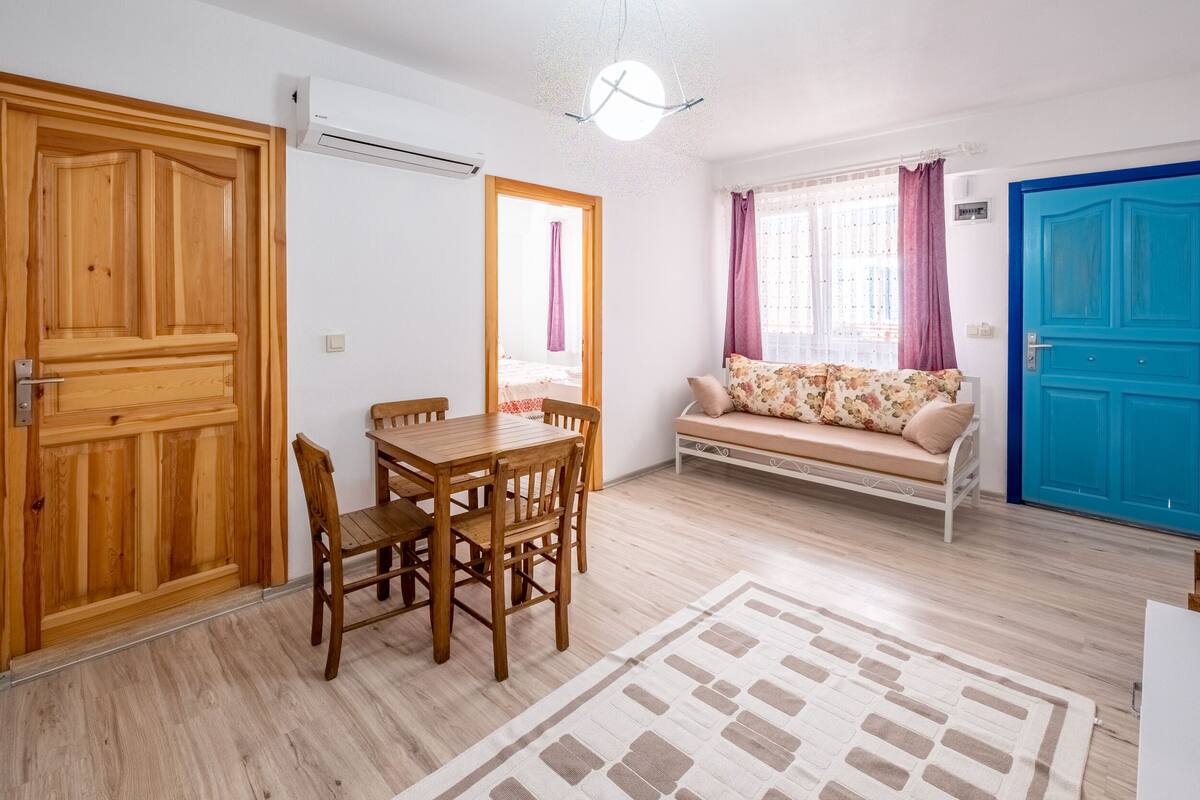 Ein Zimmer mit einem Holzespeisetisch und -stühlen, einer Couch mit floralen Kissen, einer blauen Tür und einem weißen Klimagerät.