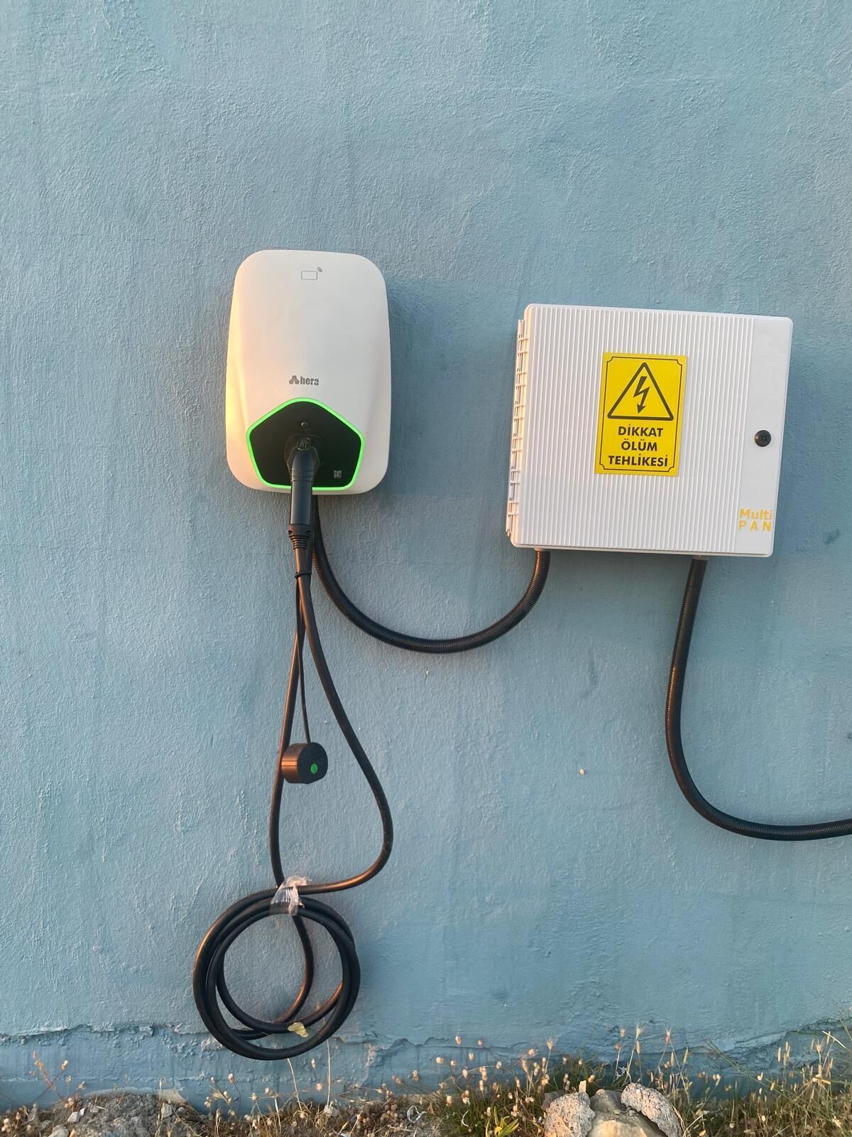 Un dispositivo bianco con una spia verde e un cavo collegato a un contenitore metallico con un cartello di avvertimento giallo.