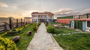 Ein zweistöckiges Haus mit einem Kiesweg, der zum Eingang führt, umgeben von einem eingezäunten Garten mit Grünflächen und einem Berg im Hintergrund.