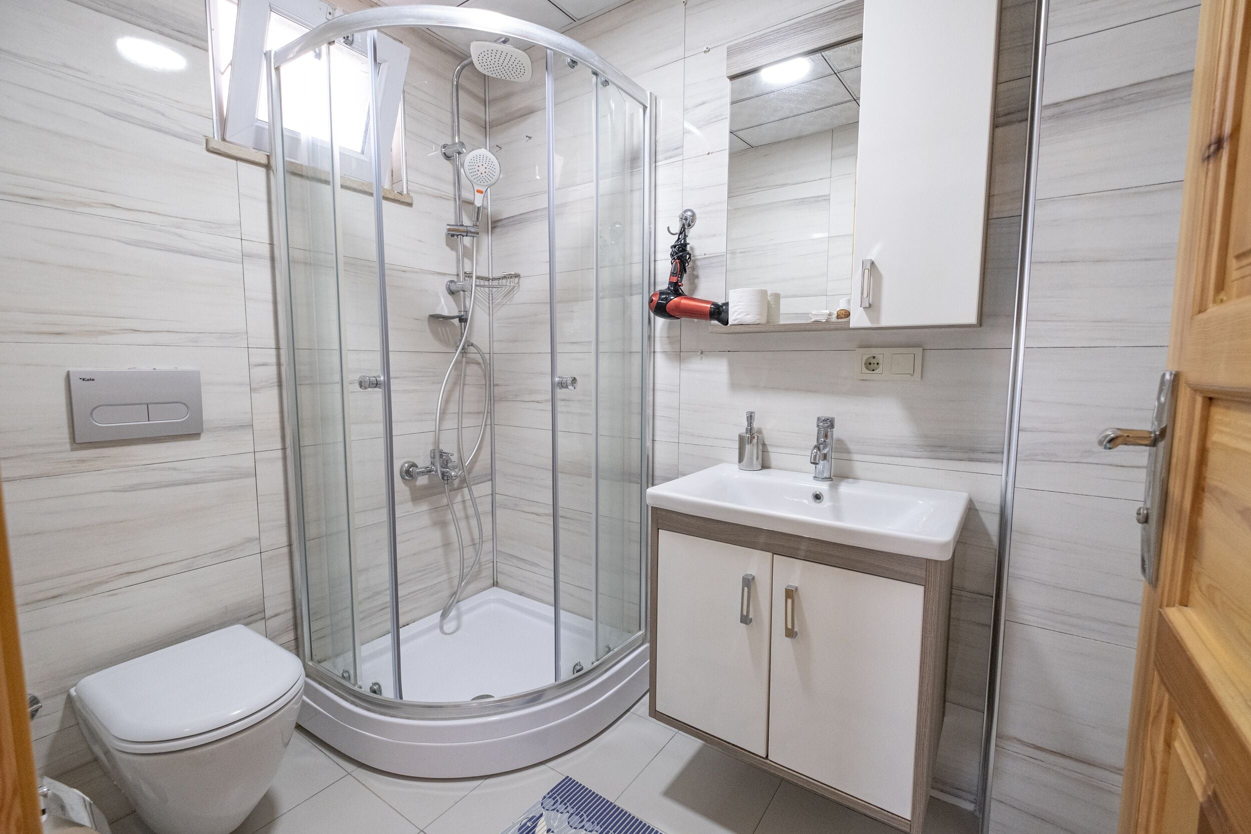 Un baño moderno con ducha de vidrio, inodoro blanco, lavabo con espejo y un mueble de madera.