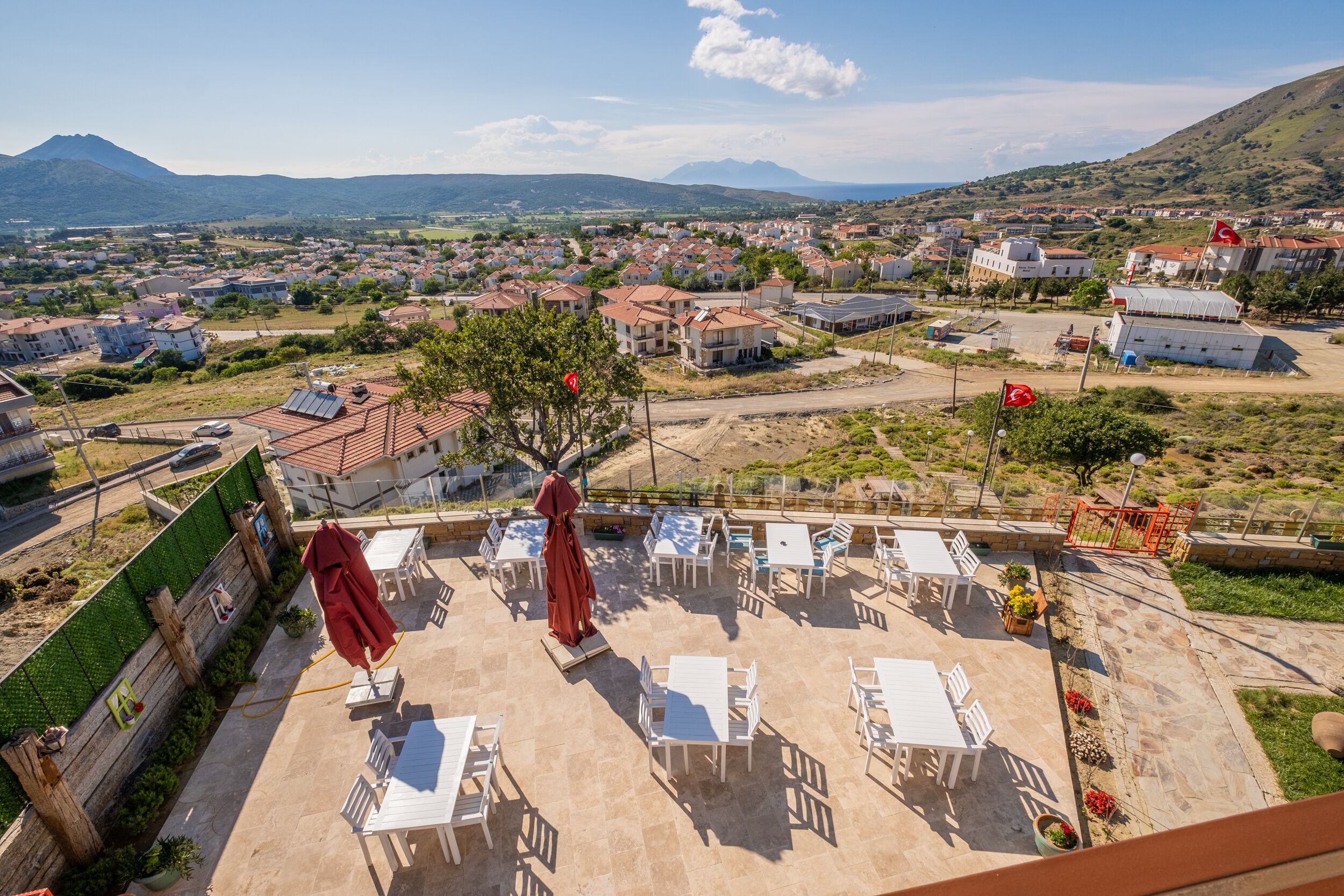 An outdoor patio with white tables and red umbrellas, overlooking a residential area with houses and a mountain in the background.