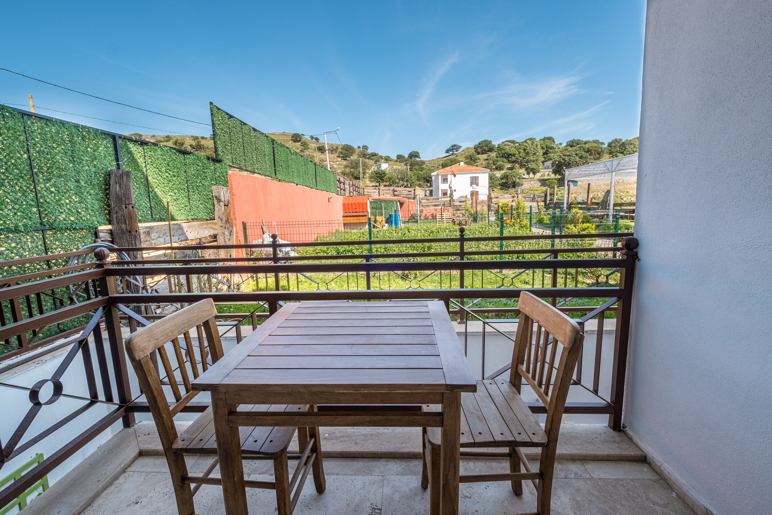 Un balcón con una mesa y sillas de madera, vista a un muro verde y edificios al fondo.