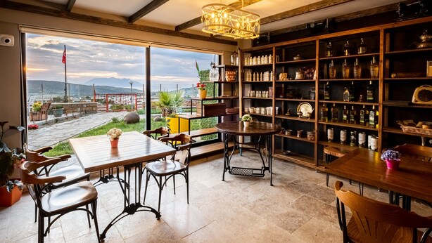 Ein geräumiges Zimmer mit großem Fenster und Blick auf Garten und Berge. Es beinhaltet einen Holztisch mit Stühlen, ein Bücherregal voller Flaschen und eine Hängeleuchte.