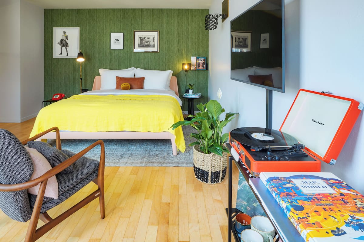 A bedroom with a bed, a chair, a plant, a record player, and a book on a table.