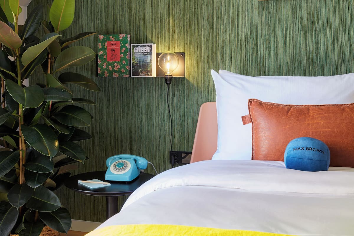 A neatly made bed with a white and yellow bedspread, a brown pillow, and a blue sports ball. A vintage telephone is on a small table beside the bed. The wall behind features a green textured wallpaper and two framed pictures.