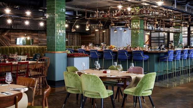A restaurant interior with wooden tables, green upholstered chairs, and a bar area with blue chairs. - Max Brown Hotel 7th District, part of Sircle Collection (Vienna)