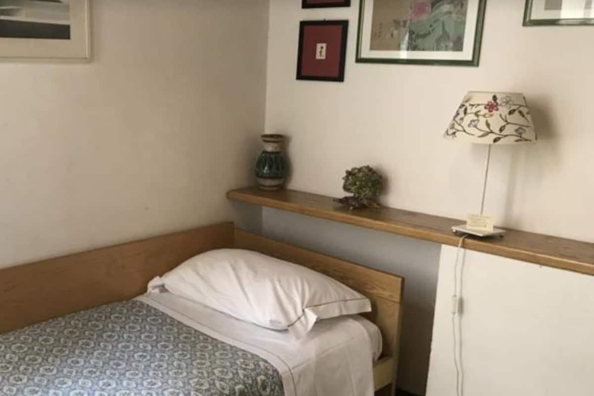 Una habitación pequeña y ordenada con una cama individual, una mesita de noche con lámpara, y un estante con objetos decorativos.