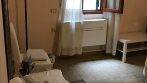 Une petite pièce, peu meublée, avec un radiateur, une chaise, une table et une fenêtre avec des rideaux.