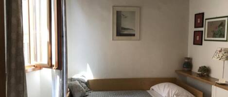 A bedroom with a bed, a window, a wall-mounted shelf with a lamp, and framed pictures.