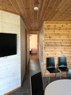 Ein Holzinnenraum mit an der Wand montiertem Fernseher, einem zur Kammer führenden Gang und zwei schwarzen Stühlen neben einem Tisch.