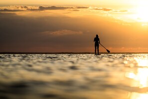 Eine Silhouette einer Person, die im Sonnenuntergang auf einem Paddleboard fährt, mit ruhiger See und bewölktem Himmel im Hintergrund.