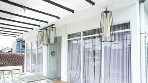 Un espacio exterior moderno con un mobiliario de madera, mesas y sillas, ventanas amplias con cortinas e iluminación colgante.