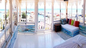 A room with a large bay window overlooking a marina, a sofa with colorful cushions, and a small table. - Denizkizi Motel (Ayvacik)