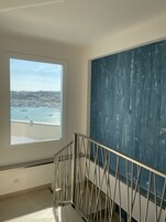 Una habitación con un ventanal grande que da a un puerto, una escalera con barandilla de metal y un cuadro abstracto azul en la pared.