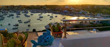 Un pueblo costero con un puerto deportivo lleno de barcos, un atardecer de fondo y esculturas decorativas de peces en el balcón.