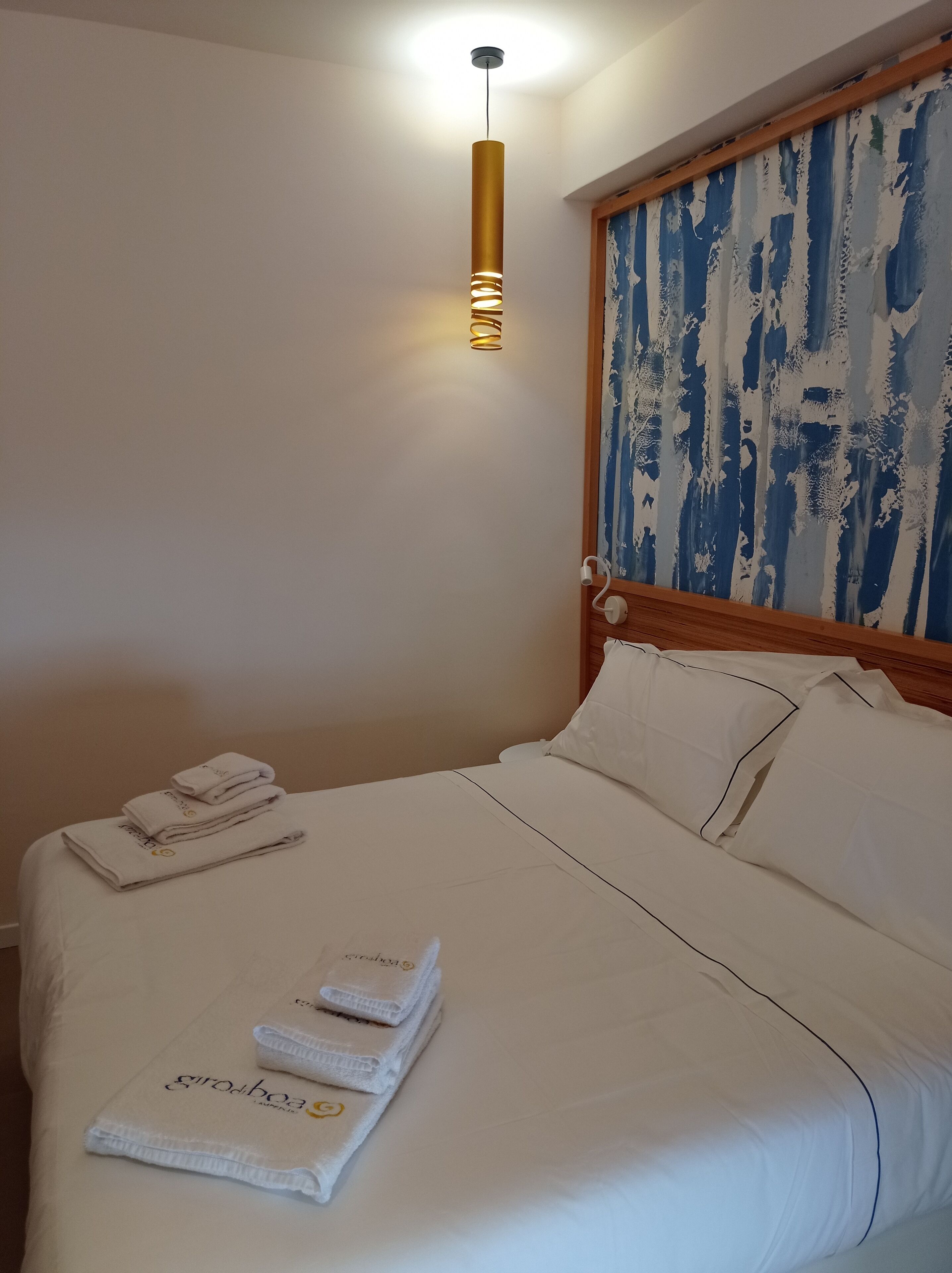 Un lit bien fait, avec du linge de lit blanc et des serviettes pliées, un luminaire suspendu moderne et un tableau abstrait aux couleurs bleu et blanc accroché au mur.