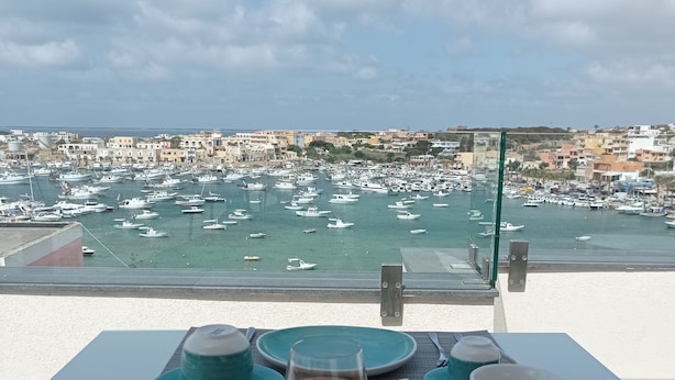 Ein Tisch, gedeckt mit Croissants, Orangensaft und Kaffee, mit Blick auf einen Yachthafen mit Booten und Gebäuden.