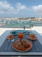 Una mesa con cruasanes, zumo de naranja y café, con vistas a un puerto deportivo con barcos y edificios.