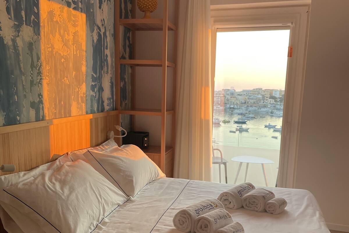 A hotel room with a bed, a wooden shelf with decorative items, a view of a harbor with boats, and a white chair by the window.