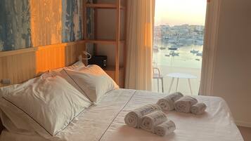 Habitación de hotel con una cama, un estante de madera con objetos decorativos, vista a un puerto con barcos y una silla blanca junto a la ventana.