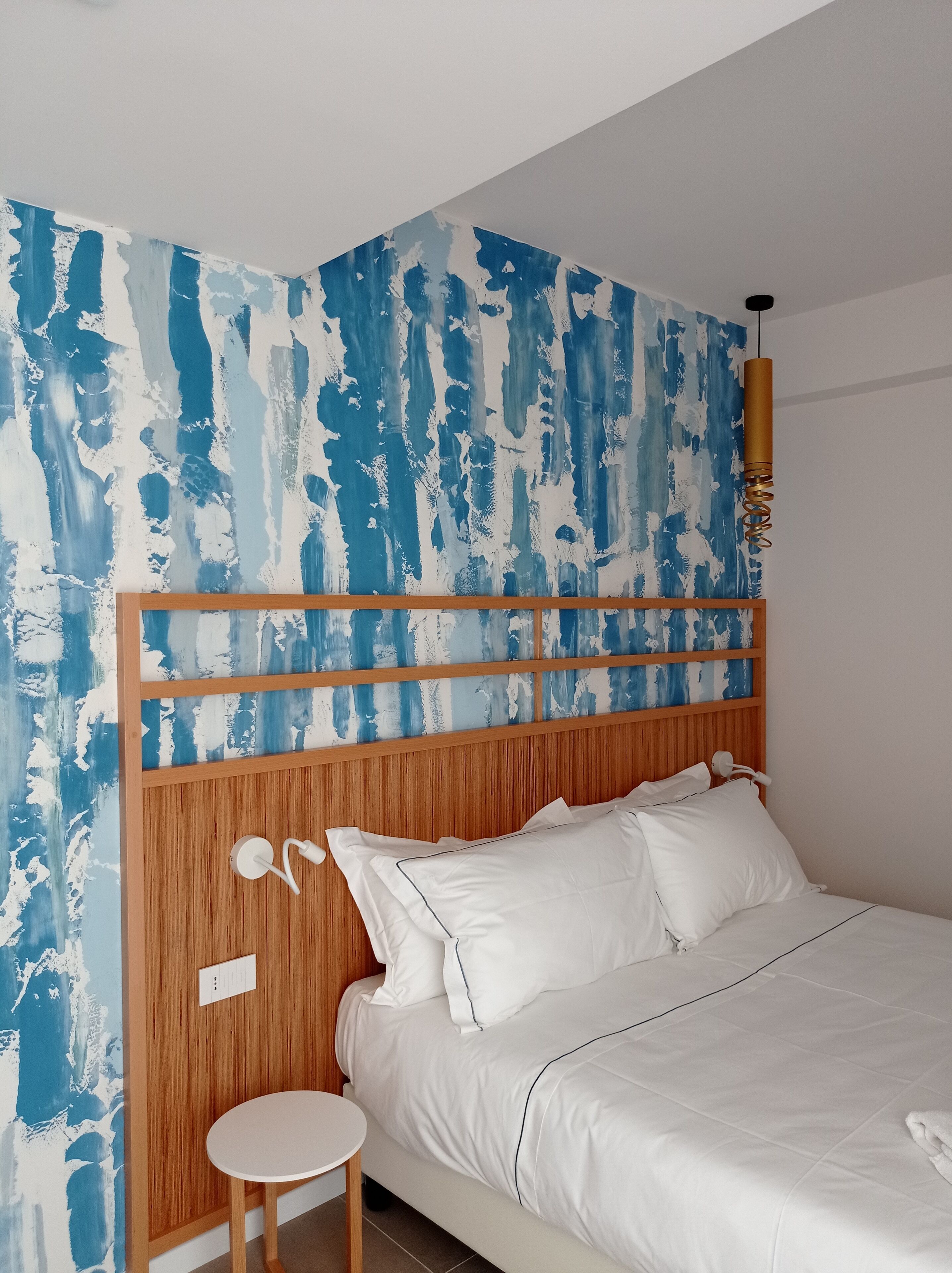 A bedroom with a wooden headboard, white bedding, and a blue abstract wall mural.