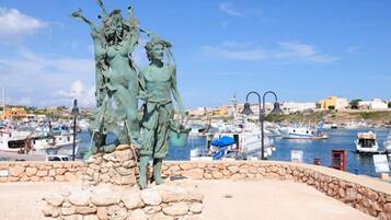 Uma estátua de duas figuras numa área delimitada por pedra, com um porto e barcos ao fundo.