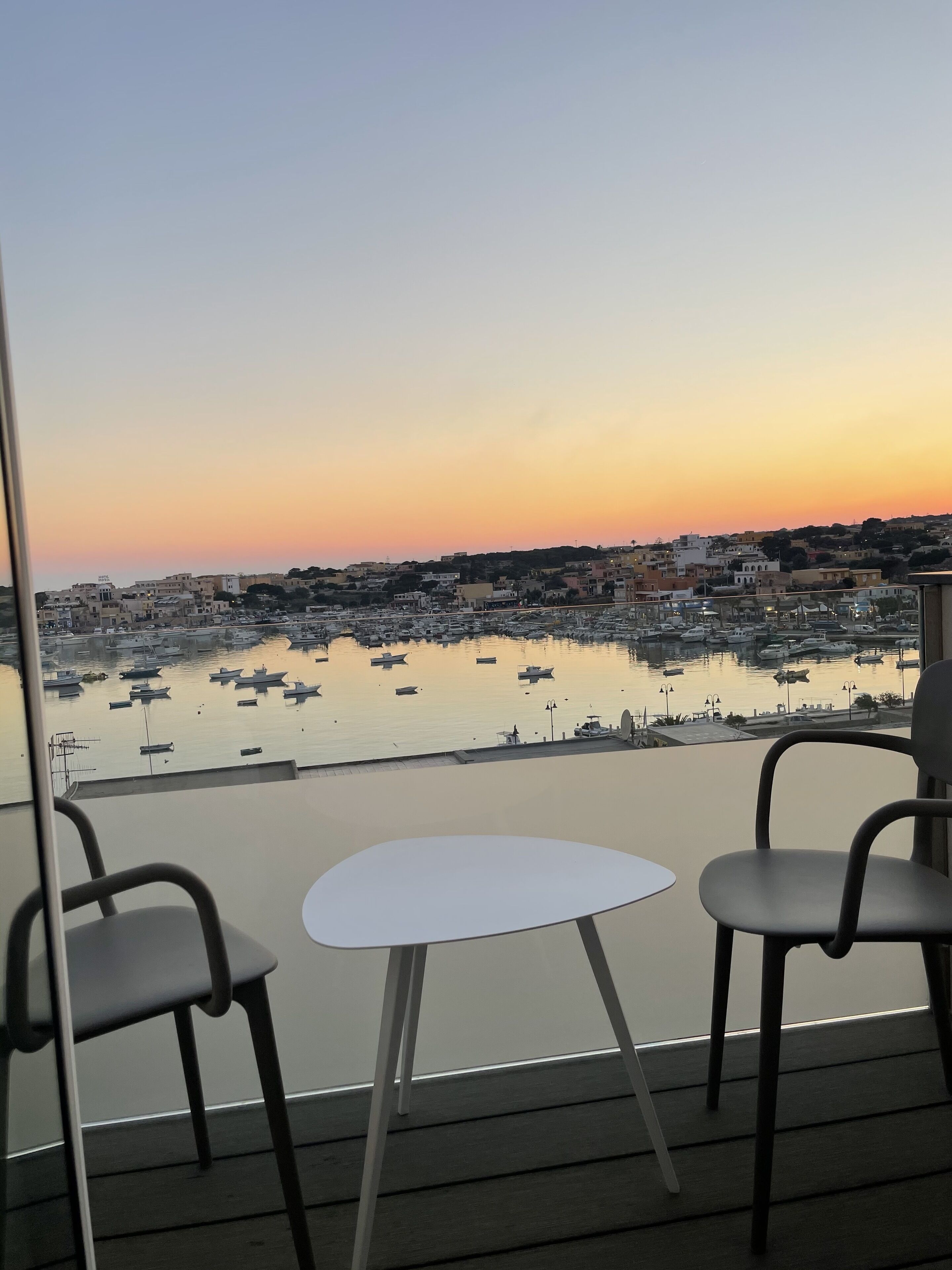 A balcony with two chairs and a small table, overlooking a marina with boats and a cityscape during sunset.