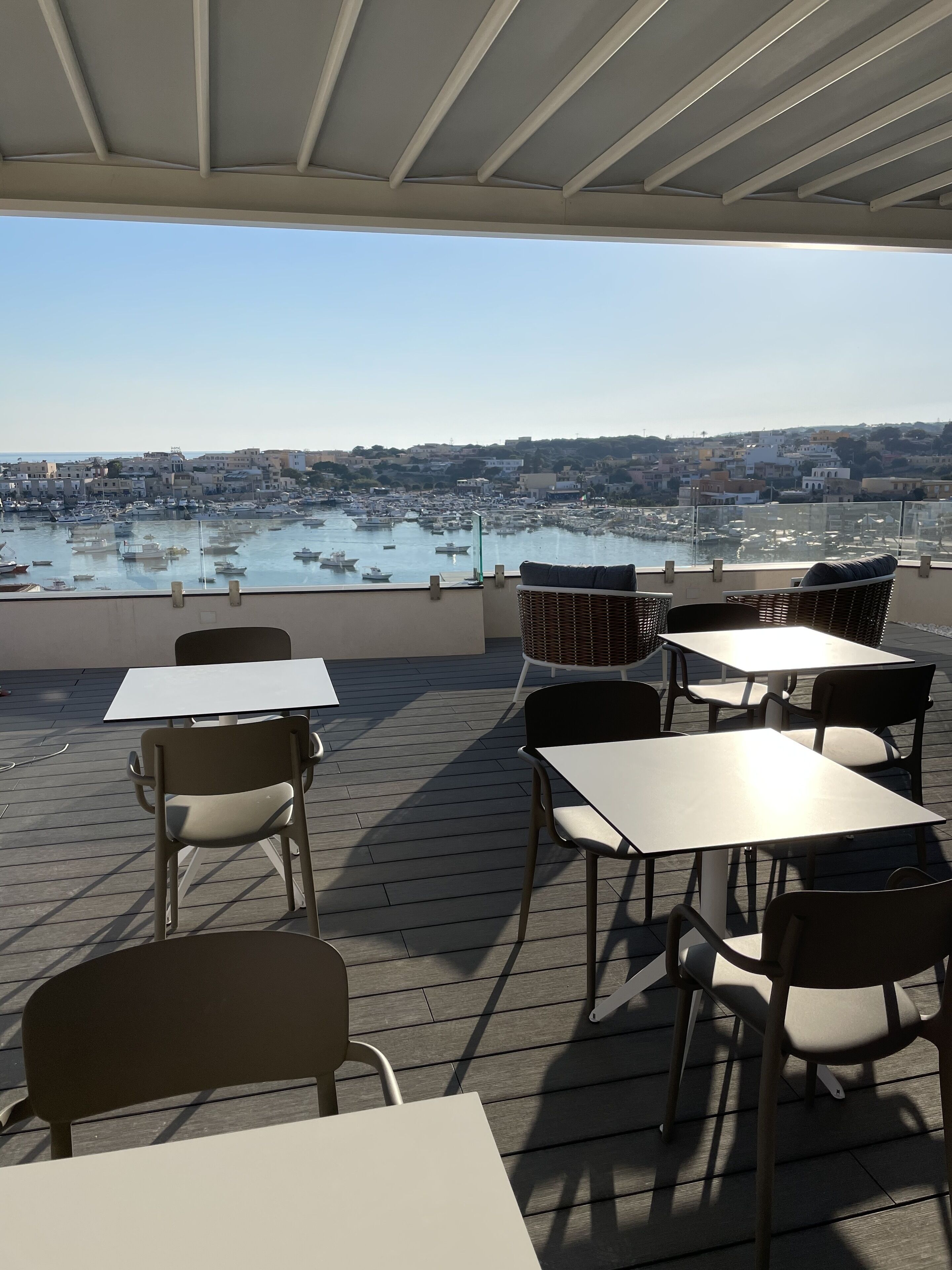 An outdoor seating area with tables and chairs, overlooking a marina with boats and a coastal town in the background.